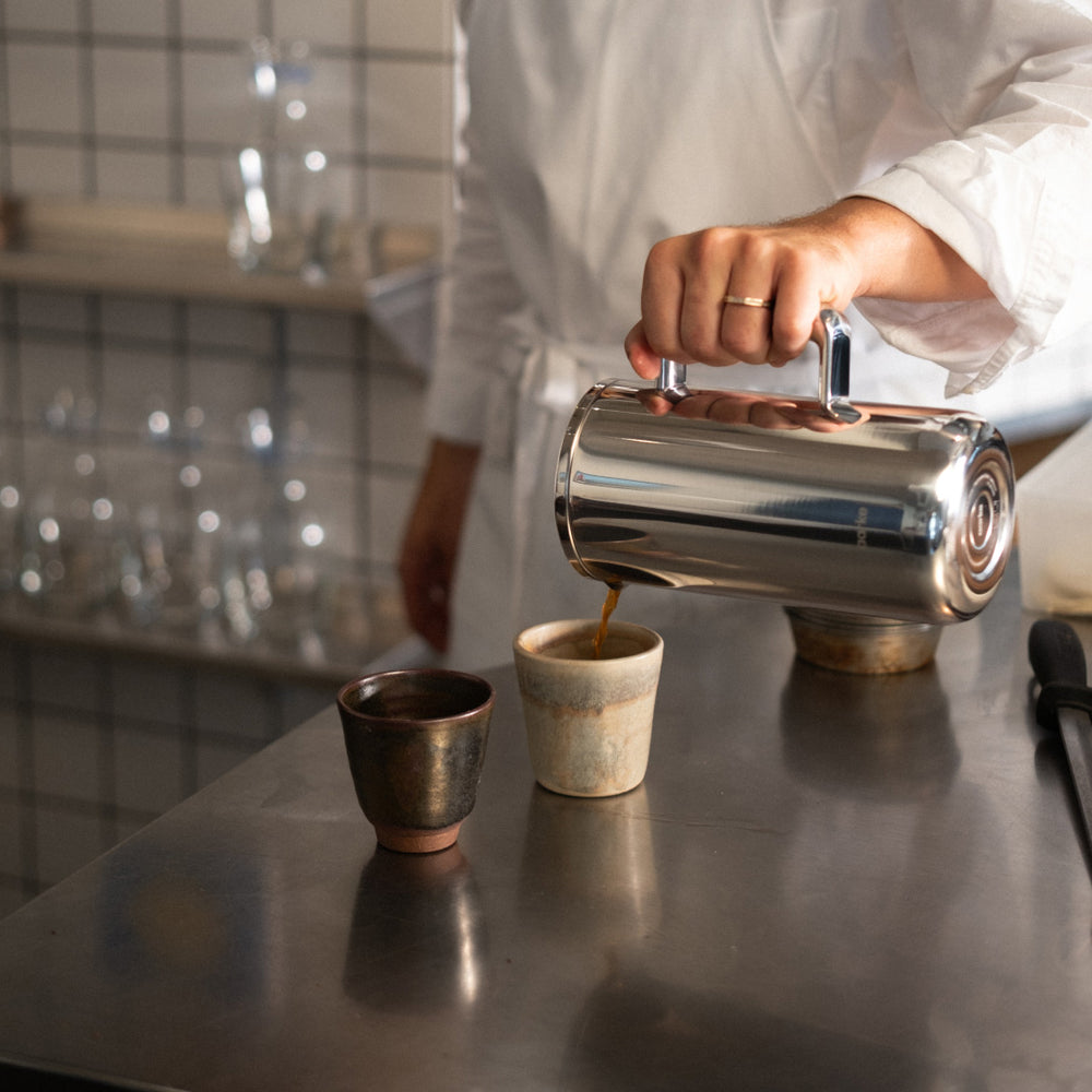 Person pouring coffee from a coffee press into a ceramic cup on a reflective surface.