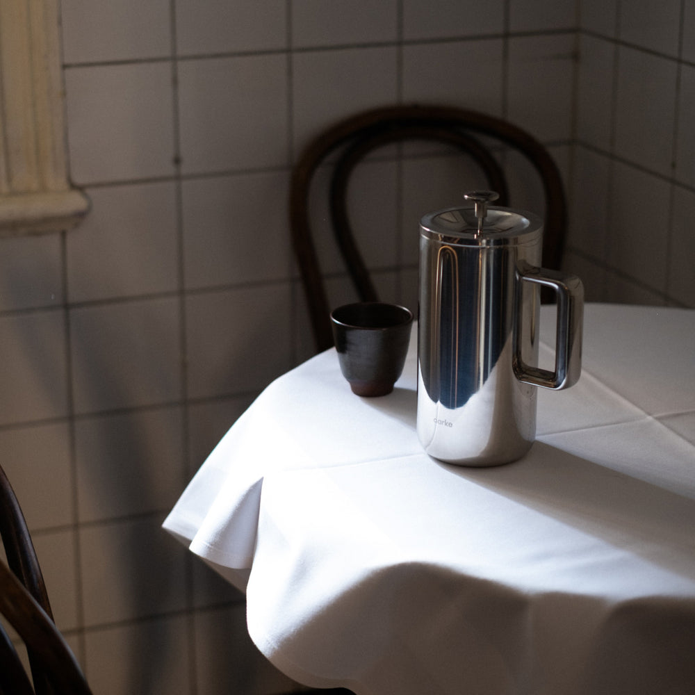 Steel Coffee Press on a white tablecloth with a tiled wall background