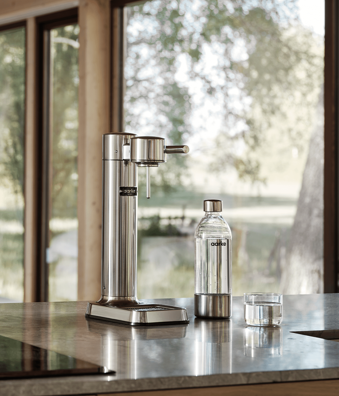 Aarke Carbonator 3 in stainless steel on a kitchen island with an Aarke PET bottle filled with sparkling water and a nesting glass beside it.