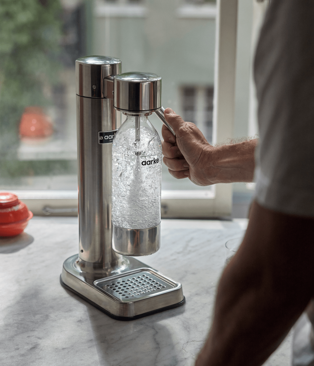 Person using the Aarke Carbonator 3 in stainless steel, carbonating water in a reusable pet water bottle on a marble countertop.