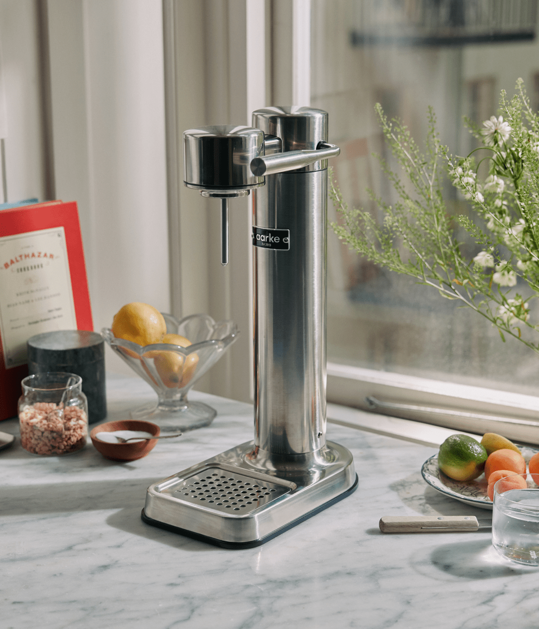 Aarke Carbonator 3 in stainless steel on a marble countertop, styled with fresh fruit and ingredients for sparkling water.