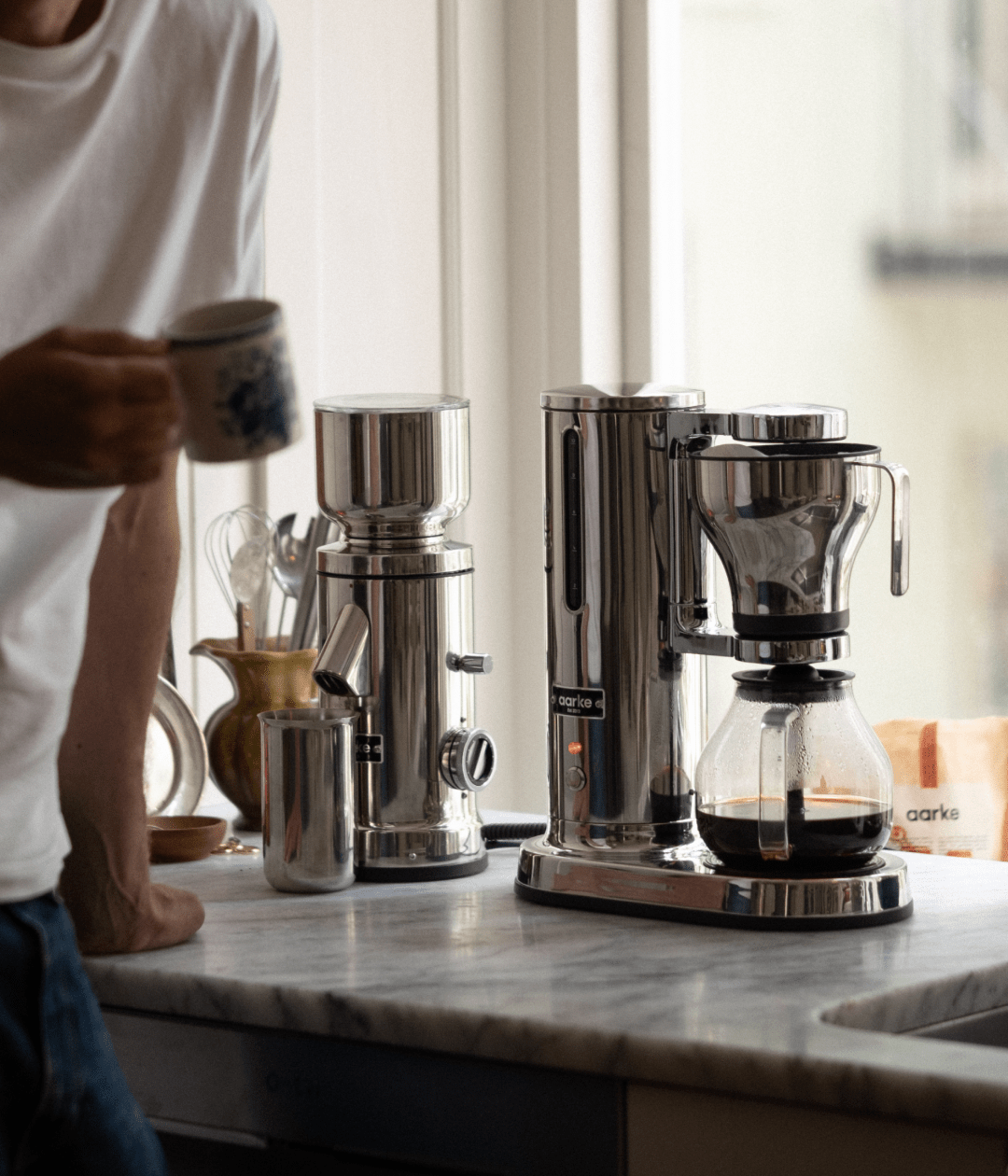 Person holding a coffee mug beside the Aarke steel coffee maker and grinder, with freshly brewed coffee in the glass carafe.