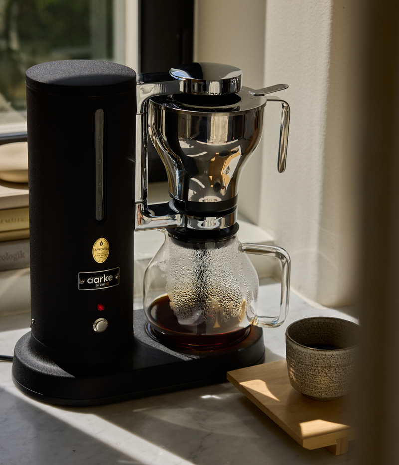 Aarke Coffee Maker Matte Black with a glass carafe on a marble surface in sunlight.