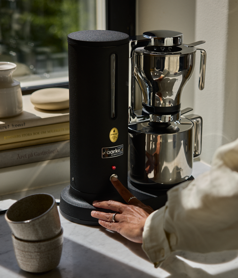 Aarke Coffee Maker in Matte Black on a counter brewing directly in Thermal Jug, surrounded by books and a window in the background.