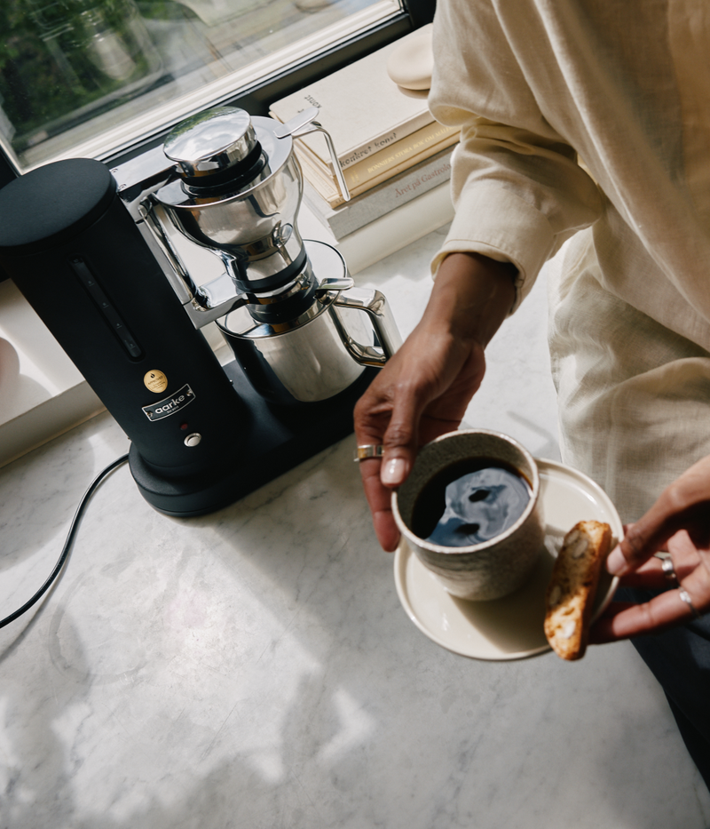Person holding a coffee cup with the Aarke Coffee Maker Matte Black on a marble surface in he background.