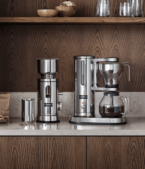 Aarke Coffee Maker and Grinder standing on kitchen countertop in a setting of dark wood. 