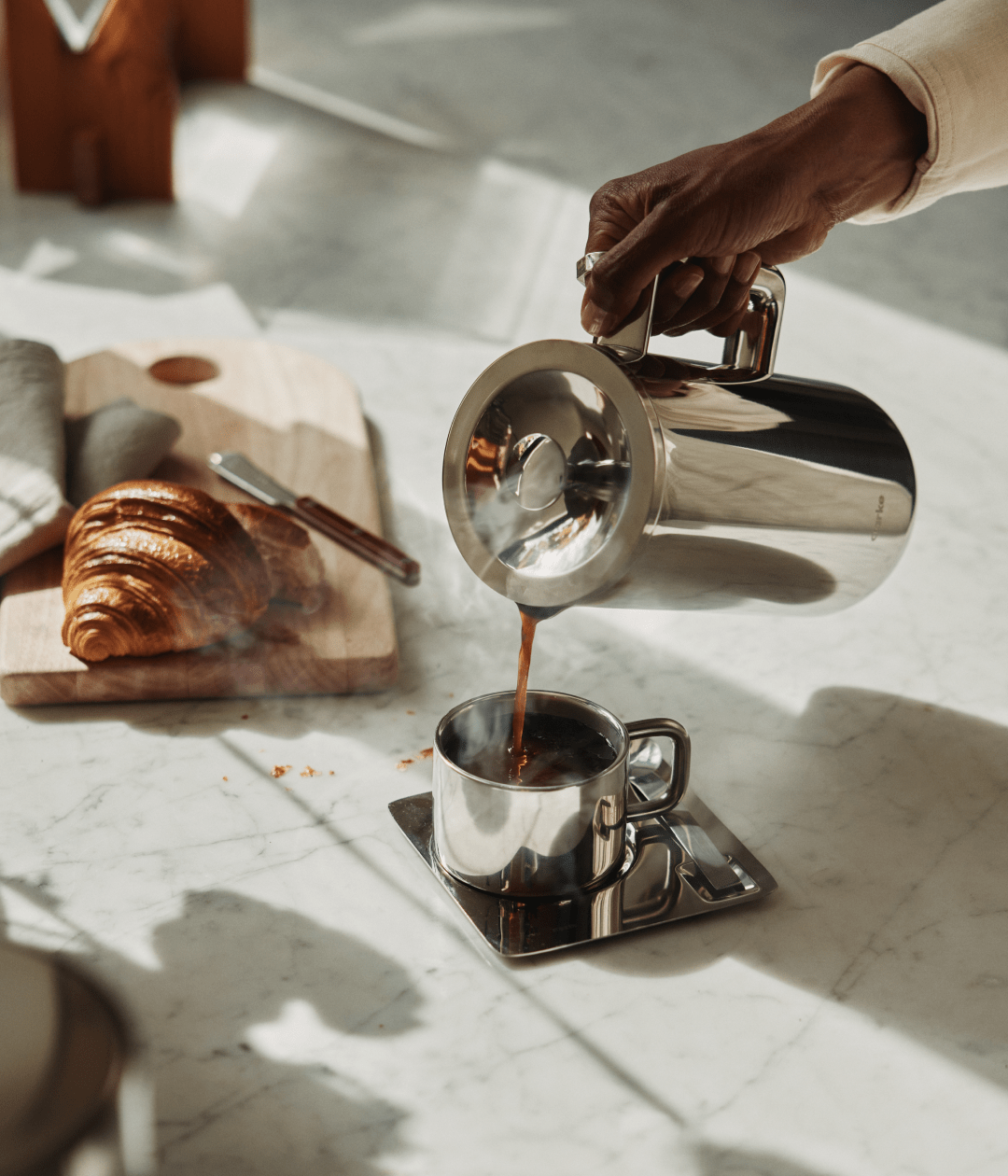 Person pouring fresh coffee from Aarke stainless steel French Press with croissant breakfast.