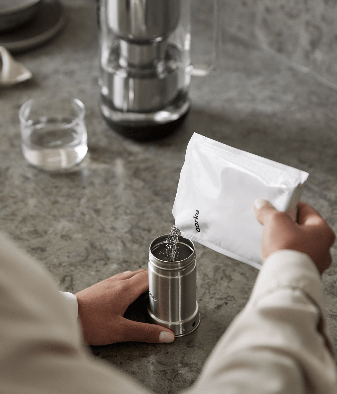 Close-up of a person pouring granules from a refill bag into a stainless steel filter cartridge.