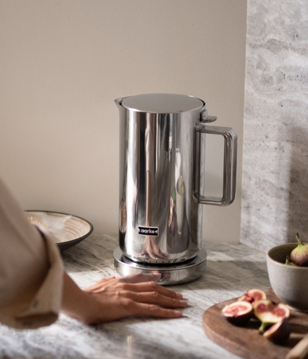 Aarke electric kettle in polished stainless steel on a marble countertop with a person’s hand.