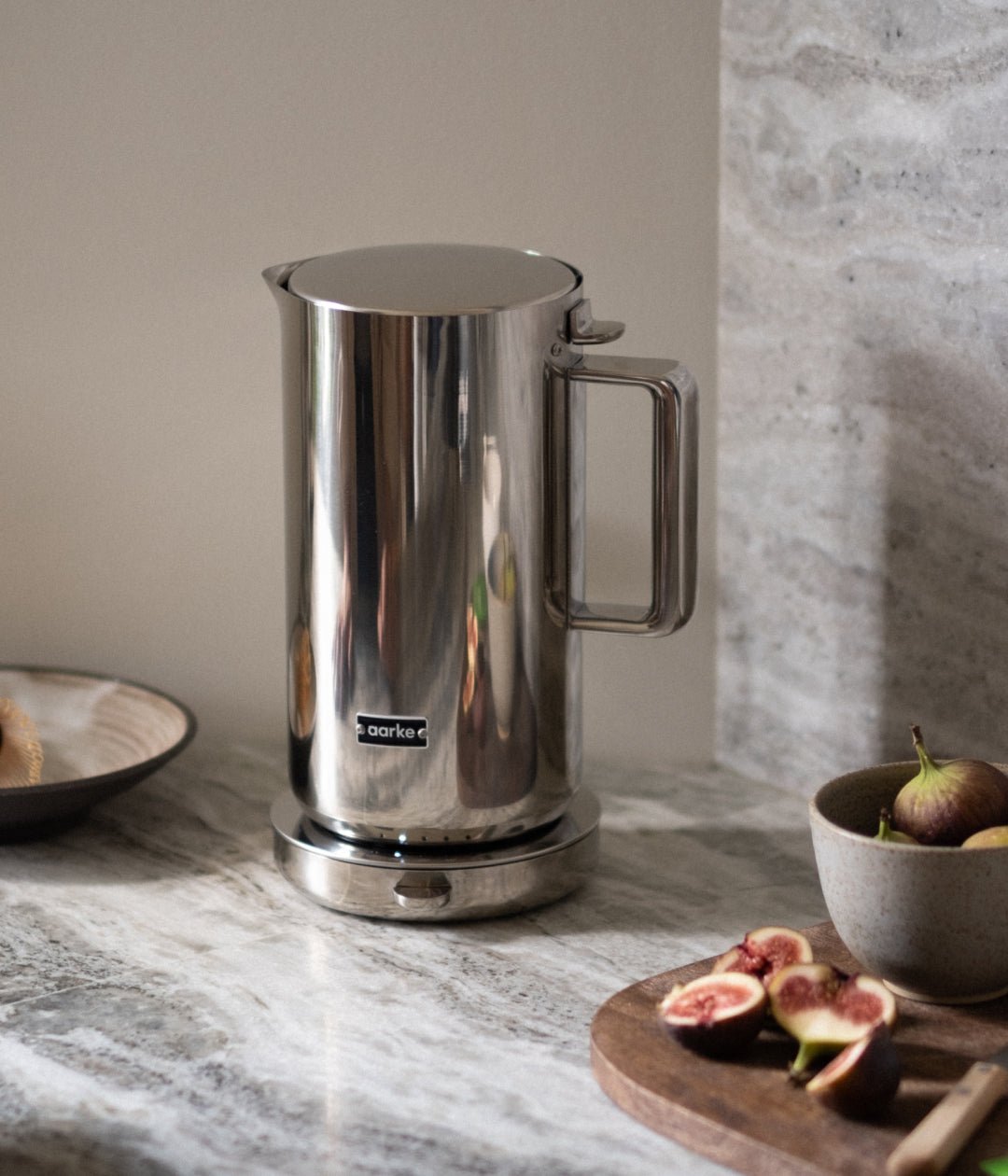 Aarke electric kettle in polished stainless steel on a marble countertop beside figs and a ceramic bowl.