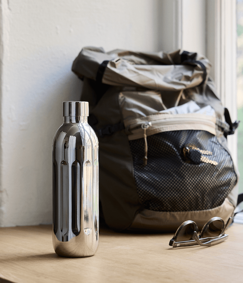 Aarke Stainless Steel Bottle Small placed on a wooden surface next to a backpack and sunglasses by a window.