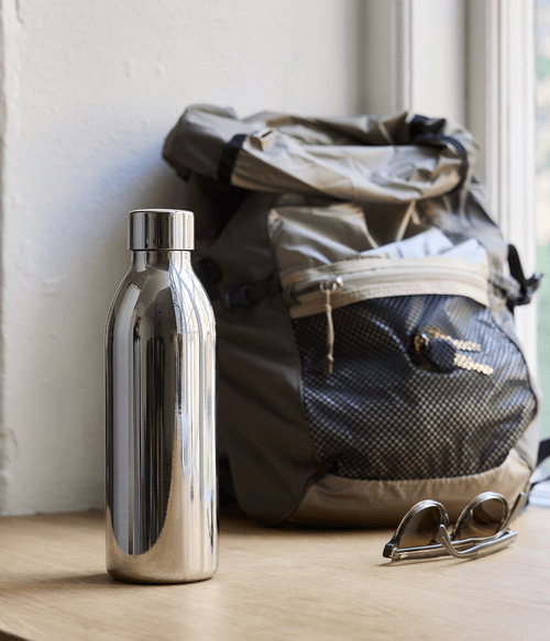 Aarke Stainless Steel Bottle Small for Carbonator 3 placed on a wooden surface next to a backpack and a pair of sunglasses by the window.