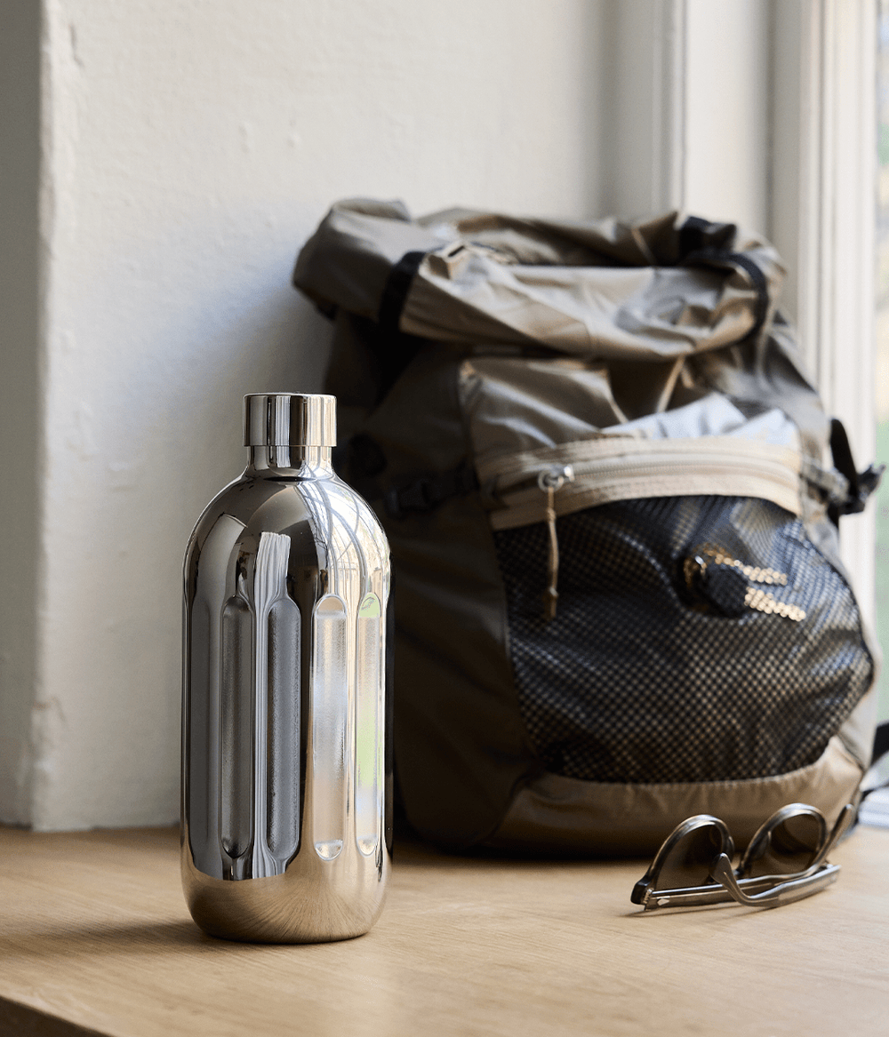 Aarke Stainless Steel Bottle placed on a wooden surface next to a backpack and sunglasses by a window.
