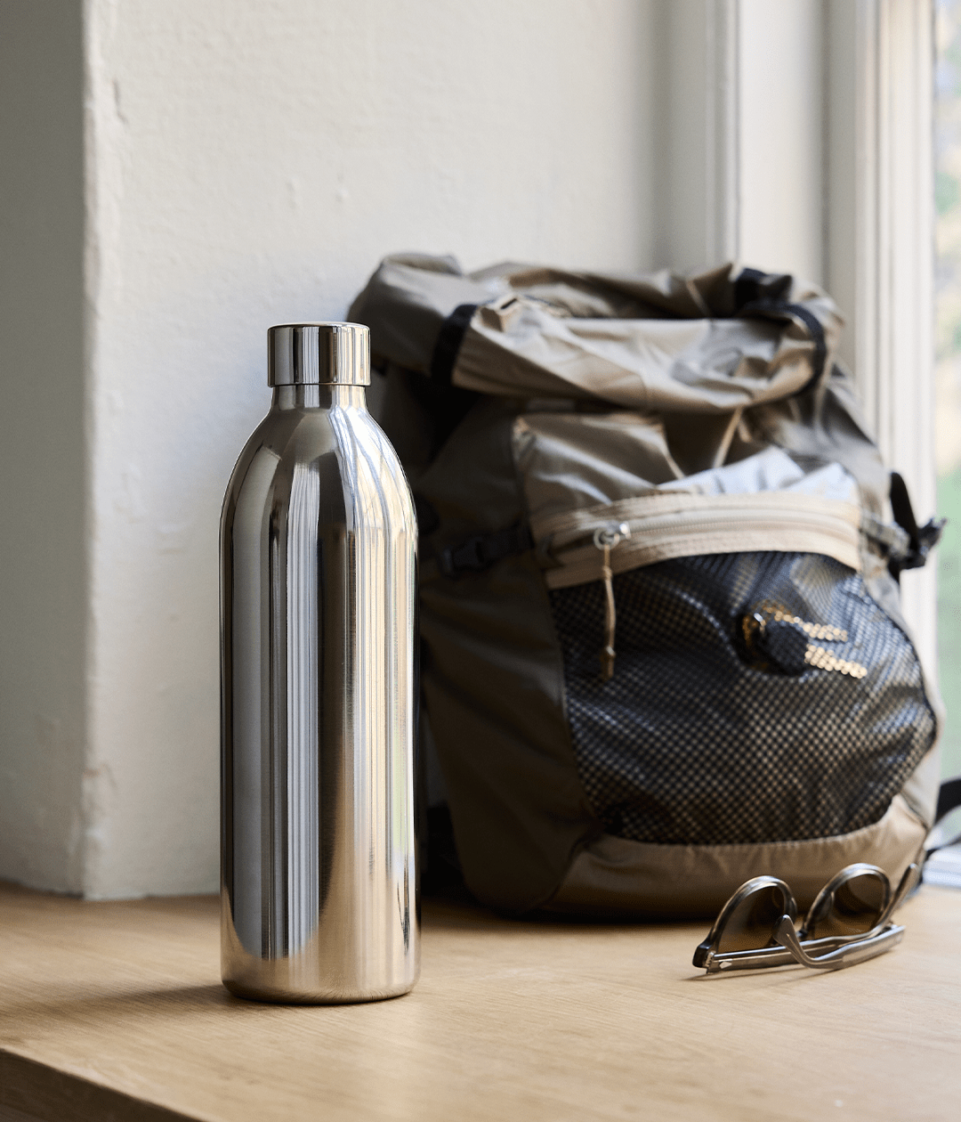 Stainless Steel Bottle for Carbonator 3 placed on a wooden surface next to a backpack and a pair of sunglasses by the window.