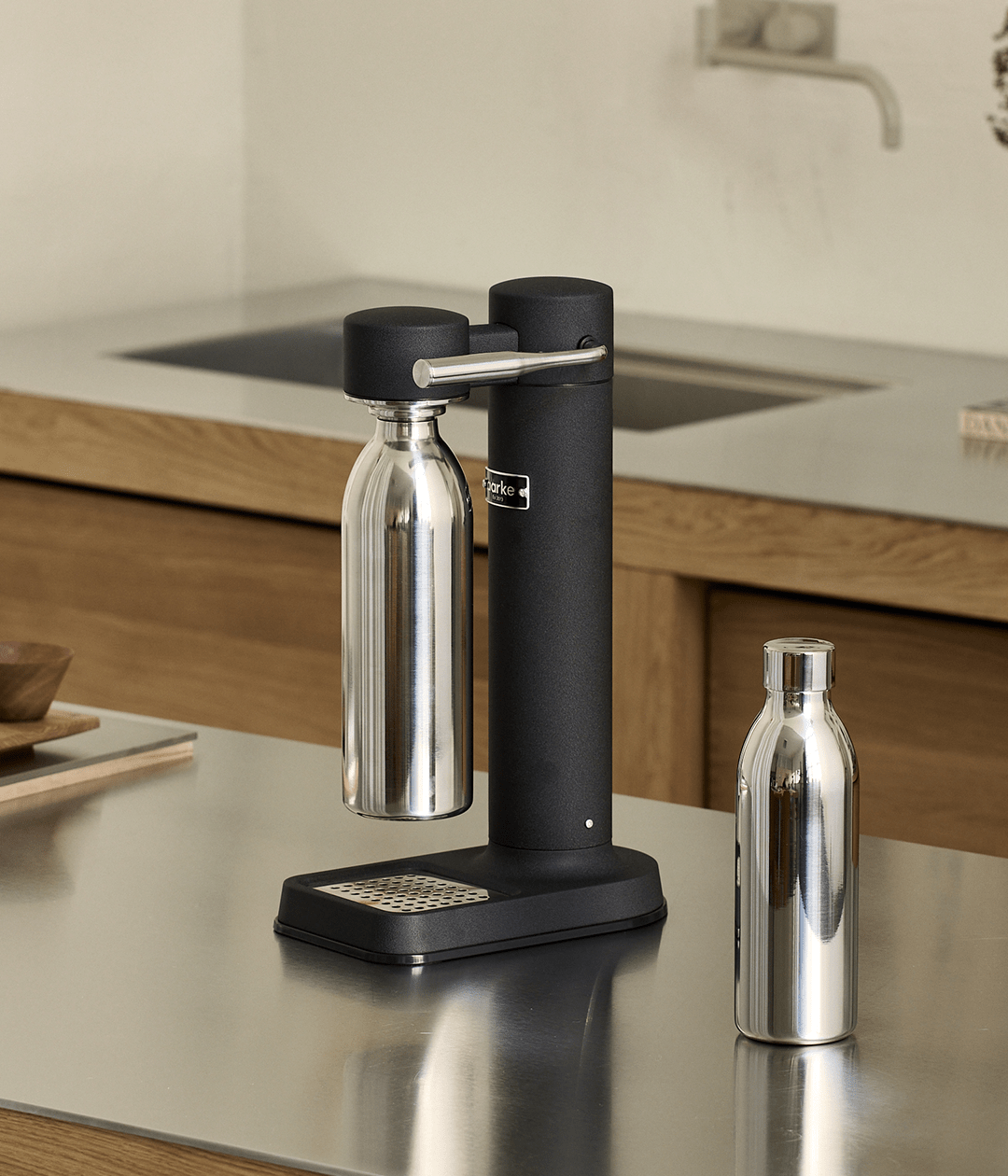 Two Aarke stainless steel bottles in a modern kitchen, one inserted into a matte black Carbonator 3, the other standing nearby.