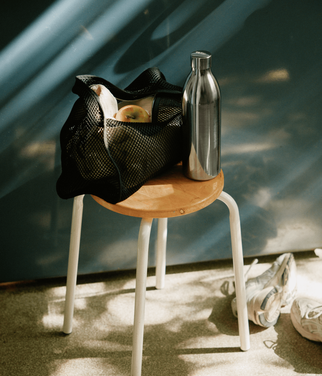 Aarke stainless steel to-go bottle placed on a stool beside a mesh bag with apples and sneakers on the floor