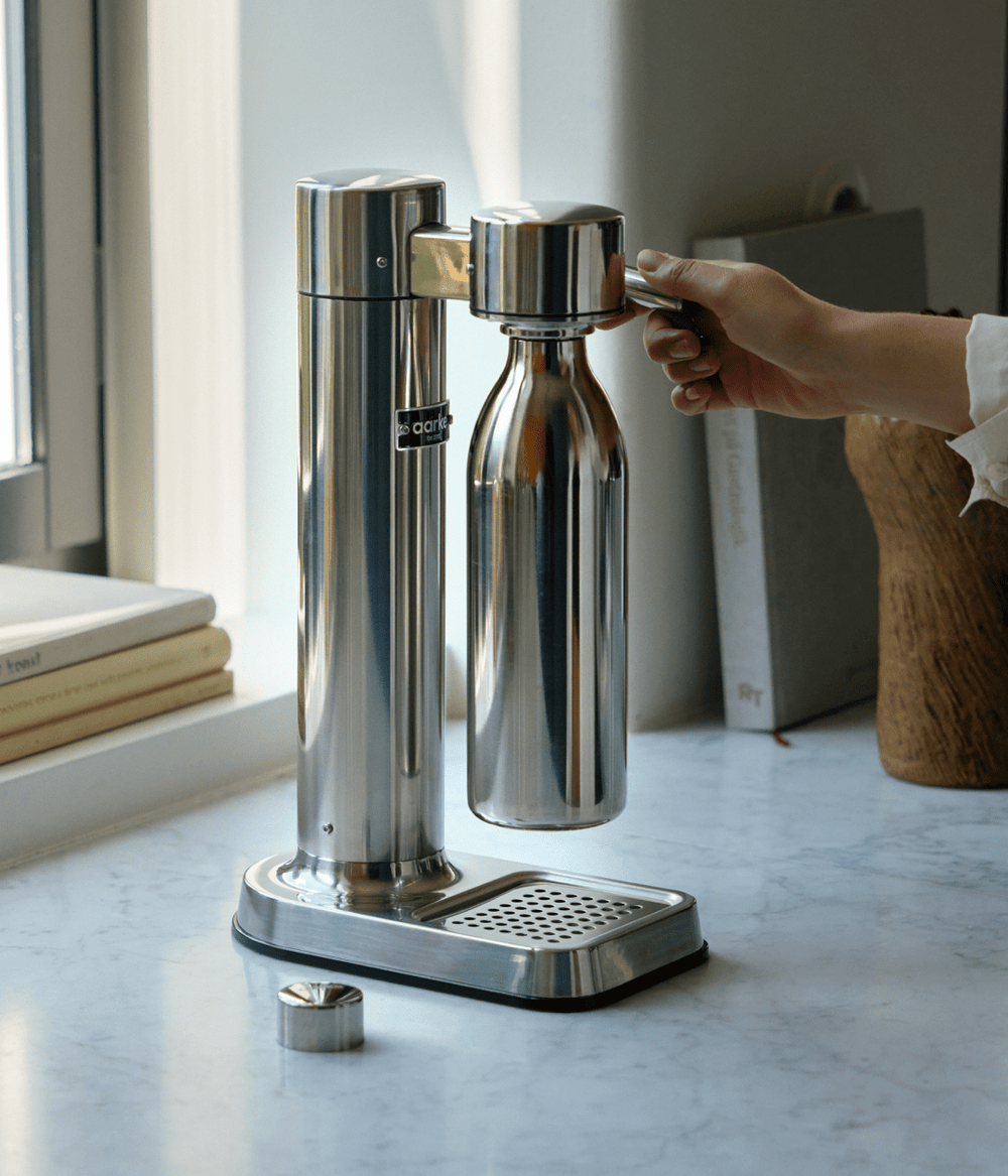 Hand pressing the lever on the Aarke Carbonator 3 in stainless steel with a matching Aarke to-go bottle attached.