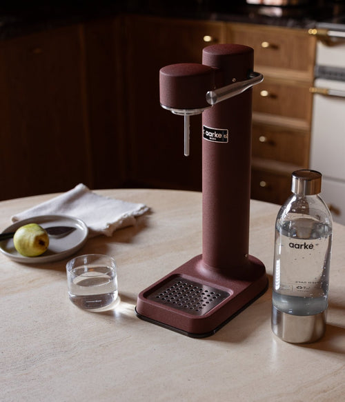 Aarke Carbonator 3 in Burgundy on a kitchen table with sparkling water and a pear.