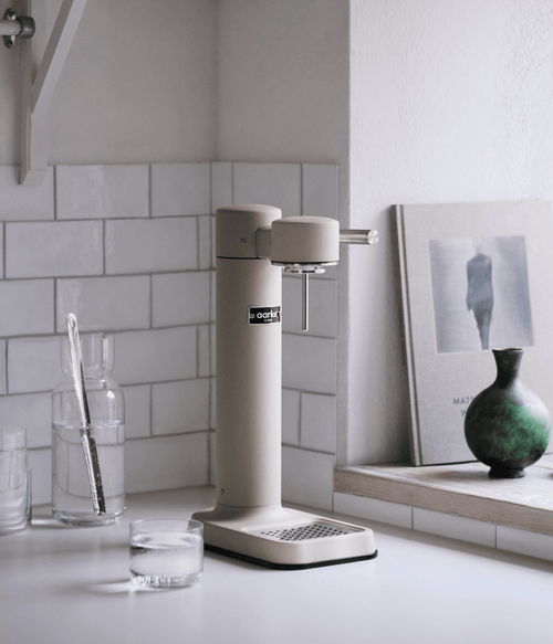 Aarke Carbonator 3 in Sand on a white kitchen counter, with decorative objects and white tiled wall in the background.