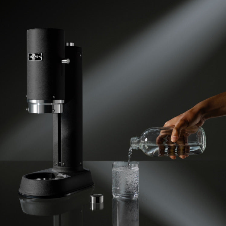 Someone pours a glass of sparkling water from the Glass Bottle sitting beside the Aarke Carbonator Pro in Matte Black.