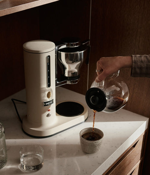 Aarke sand-colored automatic pour-over coffee maker on marble kitchen countertop as freshly brewed coffee is poured from glass carafe into ceramic cup.