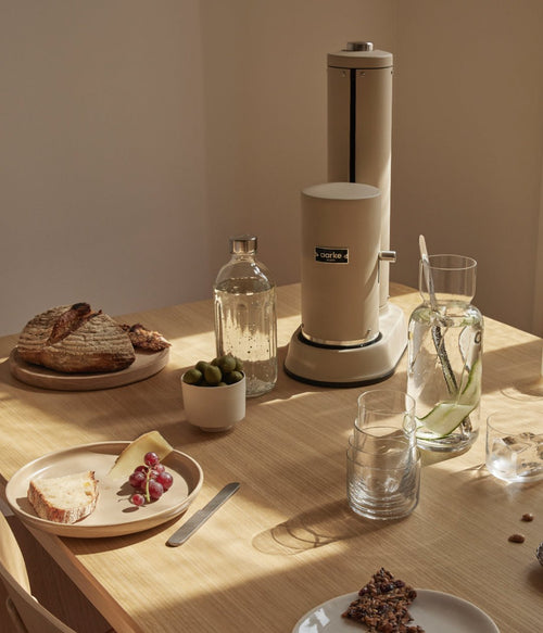 Aarke Carbonator Pro in Sand on a wooden table set for breakfast, with sparkling water and a carafe of infused water.