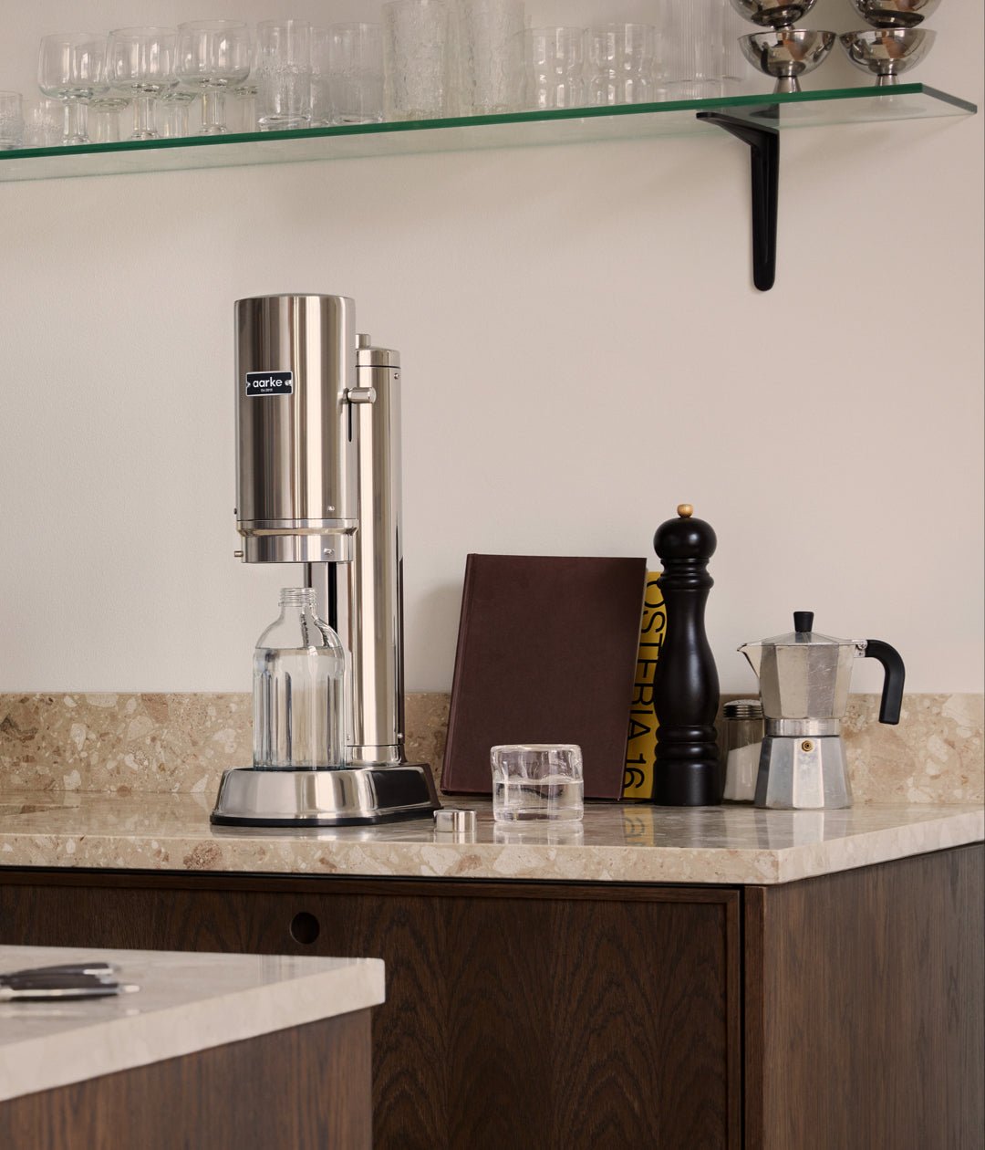 Aarke Carbonator Pro in Steel on a beige marble kitchen counter, with glassware and coffee accessories nearby.