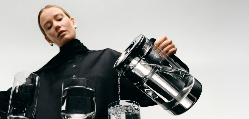 Person holding a modern water filtration pitcher with glasses filled with water on a plain background