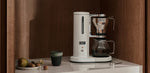 Aarke Coffee Maker in Matte White brewing filter coffee in a warm wood-paneled kitchen with a marble countertop.