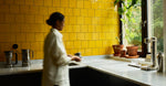 Person standing in a kitchen with yellow tiled walls and a window with plants.