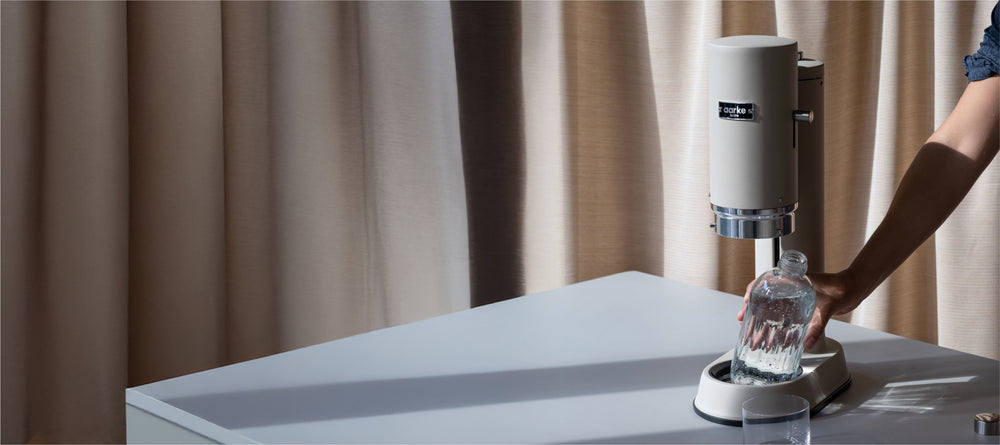 Aarke Carbonator Pro Sand on a table in front of light curtains. A person holds a glass bottle under the machine’s nozzle to carbonate water. 