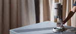 Aarke Carbonator Pro Sand on a table in front of light curtains. A person holds a glass bottle under the machine’s nozzle to carbonate water. 