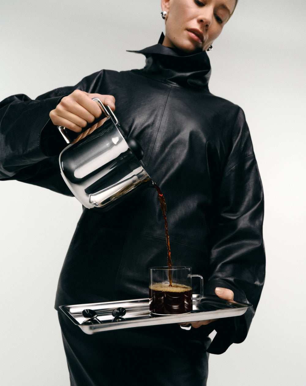 Person in a black outfit pouring coffee from a the aarke thermal jug into a glass mug on a white background