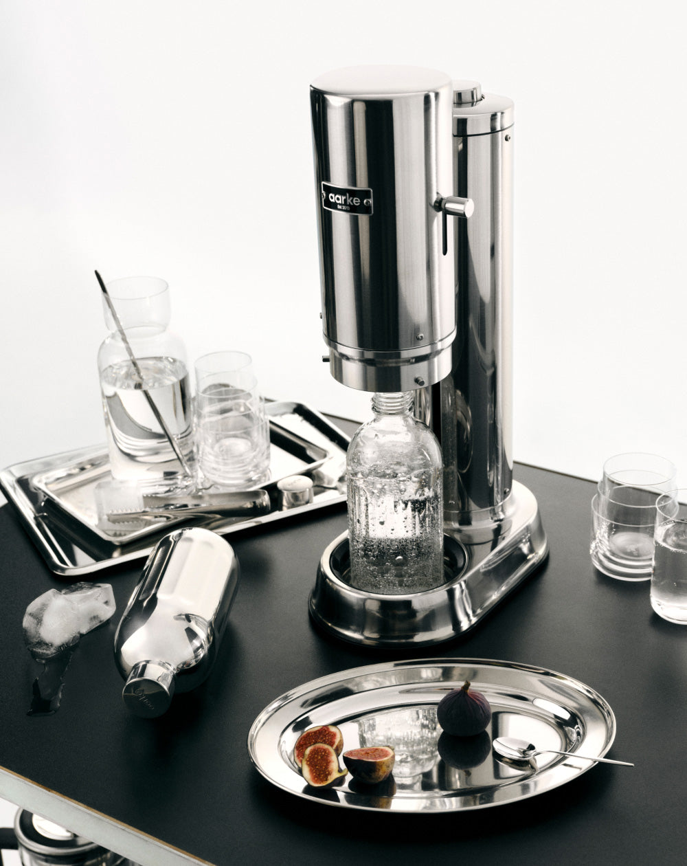 Stainless steel Carbonator Pro on a black tray with glasses and bottles.