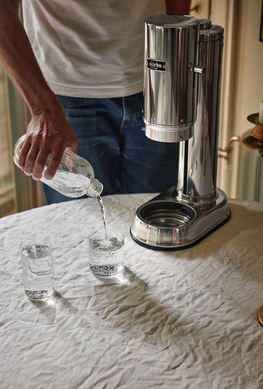 Aarke sCarbonator Pro Steel on a table with a white tablecloth. A person pours carbonated water from a glass bottle into a nesting glass, with another glass placed beside it.