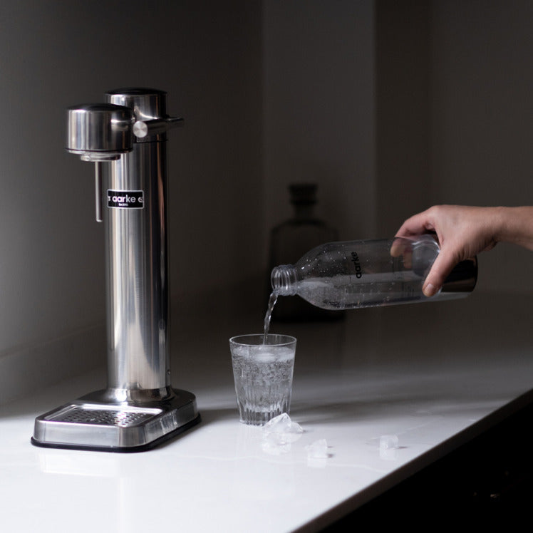 Someone pours water into a glass from the PET Bottle beside the Aarke Carbonator 3 in Stainless Steel.