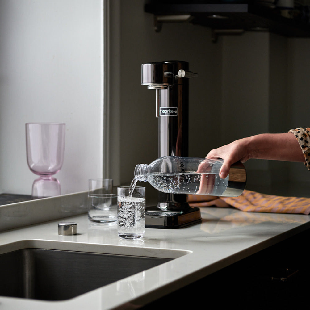 A person pours water into a glass from the Aarke PET Bottle in front of the Carbonator 3 in Black Chrome.