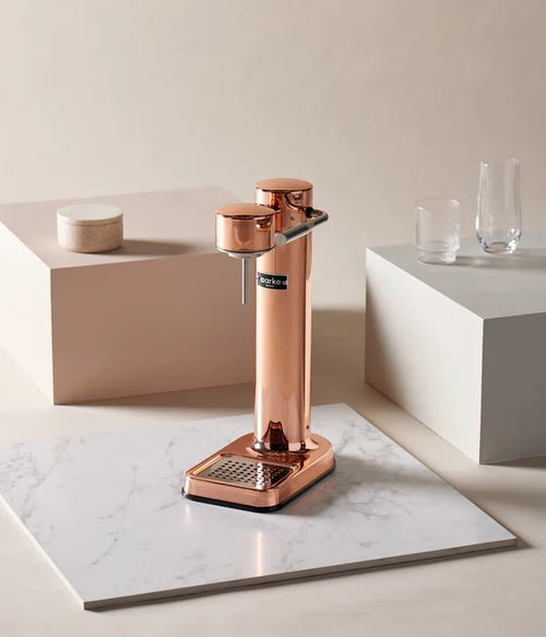 Aarke Carbonator 3 in Copper placed on a marble tile with glassware around, in a soft-toned studio setting.