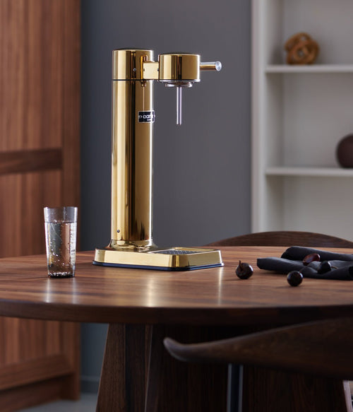 The Aarke Carbonator 3 in Gold placed on a wooden table in a modern kitchen setting, next to a glass of sparkling water.