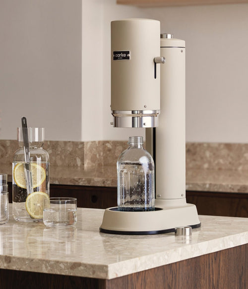 Aarke Carbonator Pro in Sand on a beige marble kitchen counter, next to a carafe of lemon water.