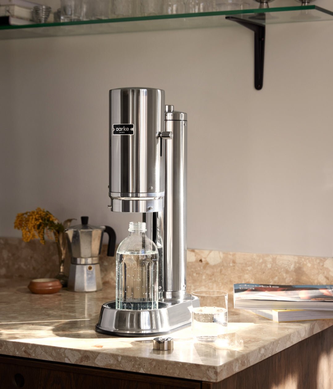 Aarke Carbonator Pro in Steel on a kitchen counter with a glass bottle inside. Sunlight highlights a nearby glass and magazines.