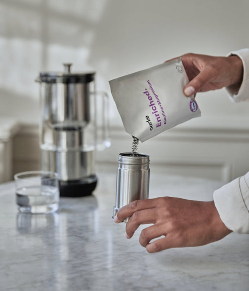 Close-up of person refilling Aarke filter cartridge with Enriched granules from refill bag on kitchen counter.