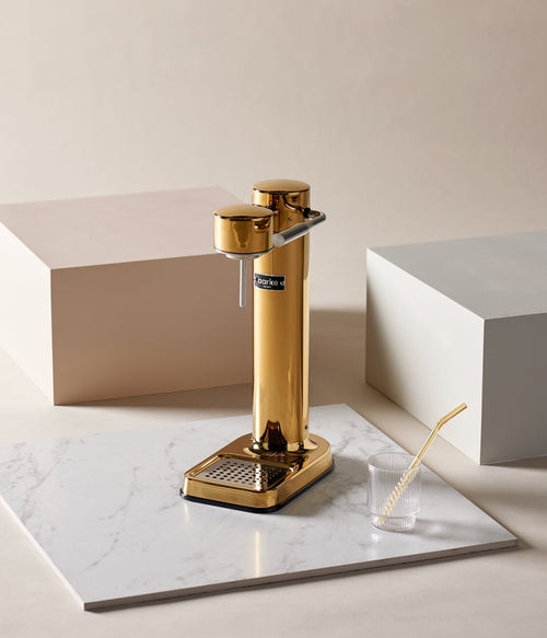 Aarke Carbonator 3 in Gold displayed on a marble surface with a ribbed glass and gold straw, set in a minimalistic studio setting.