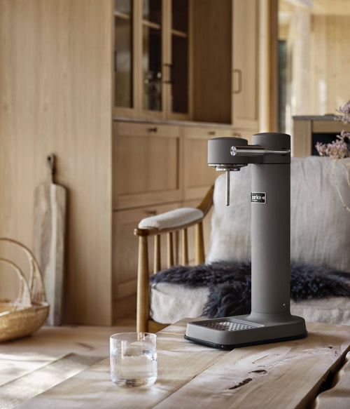Aarke Carbonator 3 in Matte Grey on a wooden table with a glass of water, in a cozy wood-accented interior.