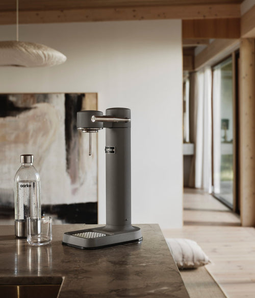 Aarke Carbonator 3 in Matte Grey finish on a stone kitchen counter, next to a PET water bottle and abstract art in the background.
