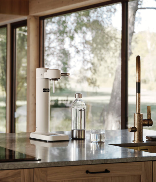 Aarke Carbonator 3 in Matte White on a stone countertop with a PET bottle, in a kitchen with large windows and a view of nature.
