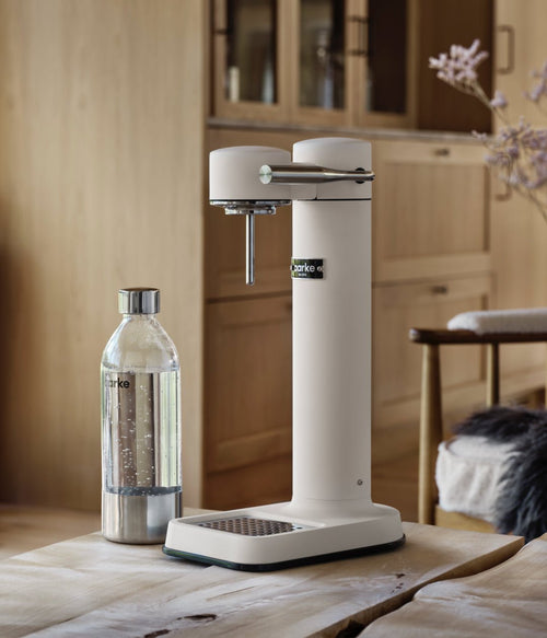 Aarke Carbonator 3 Matte White placed on a wooden table in a kitchen with wood cabinetry, next to a PET bottle filled with water.