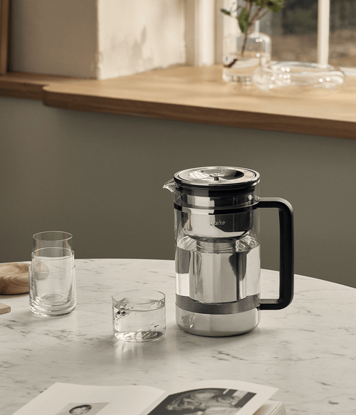 Aarke Purifier Large water filter pitcher on marble table with filtered water, drinking glasses, and magazine.