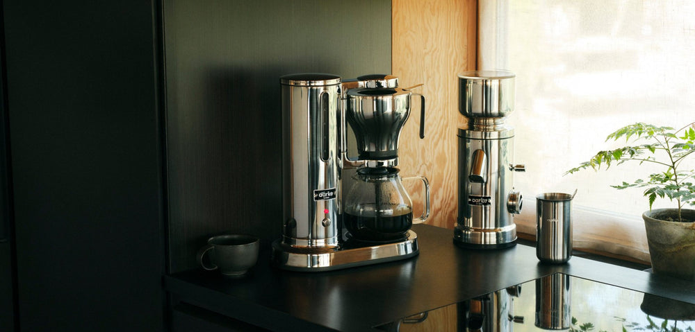 Aarke coffee maker steel and coffee grinder on a modern kitchen counter with a potted plant by the window.
