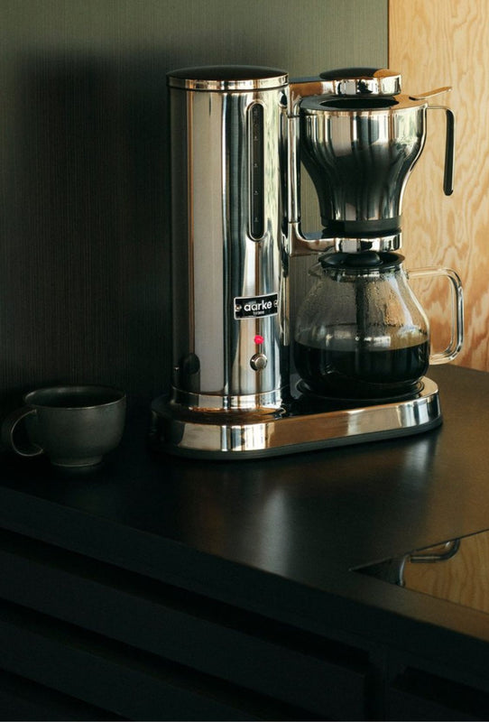 Aarke coffee maker steel on a dark kitchen counter with a ceramic cup beside it.